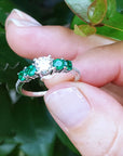 A white gold ring with emeralds held between fingers against a green leafy background.