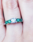 A ring with a center round diamond and side round emeralds on a finger.