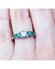 A ring with a center round diamond and side round emeralds on a finger.