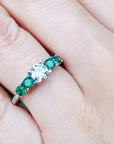 Ring with middle round diamond and side emeralds on a finger.