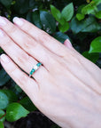 Hand wearing a ring with center diamond and side emeralds against a green leafy background.