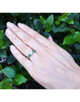 Hand wearing a ring with center diamond and side emeralds against a green leafy background.