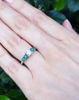 Hand wearing a ring with a center diamond and side emerald ring against a green leafy background.