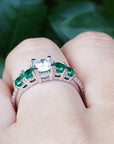 Ring with emeralds and a diamond on a finger against a green leafy background.