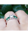 Ring with emeralds and a diamond on a finger against a green leafy background.