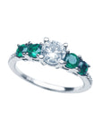 A tilted position of this ring with a round cut diamond and side emeralds displays its topside.