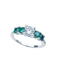 A tilted position of this ring with a round cut diamond and side emeralds displays its topside.