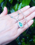 Pear cut emerald necklace in 18K white gold with double diamond halo, held in a hand.