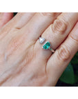 18k white gold emerald and diamond you and me ring, shown worn on a person's finger against a blurred green background.