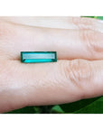 3.13 carat baguette cut Colombian emerald, sparkling bluish-green, held on a finger.