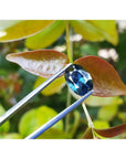 3.04 ct natural Australian deep blue oval cut sapphire held by tweezers against green leaves.