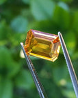 4.05 ct. vibrant intense yellow sapphire gemstone, emerald-cut, held by tweezers against a green background.