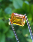 4.05 ct. vibrant intense yellow sapphire gemstone, emerald-cut, held by tweezers against a green background.