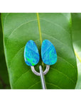 Pear-shaped, deep blue Australian doublet opals with vibrant green flashes, held by tweezers on a green leaf.