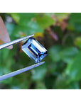 3.90 ct untreated GIA certified rectangular emerald-cut blue sapphire held by tweezers, with green foliage background.