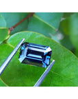 3.90 ct untreated GIA certified blue changing sapphire, rectangular emerald-cut, held by tweezers on a green leaf.