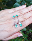 May's birthstone Love Knot emerald earrings displayed on a hand against a green leafy background
