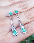 Love Knot emerald earrings held on a hand against a blurred natural background