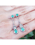 Love Knot emerald earrings held on a hand against a blurred natural background