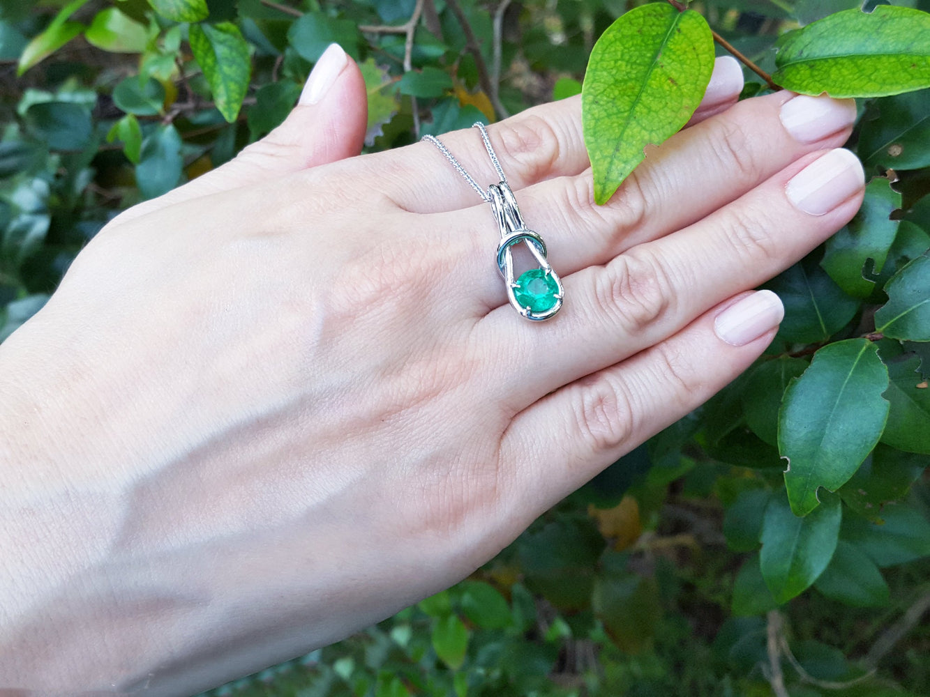 Hand displaying a necklace with an emerald pendant against a green leafy background.