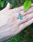 Hand displaying a necklace with an emerald pendant against a green leafy background.