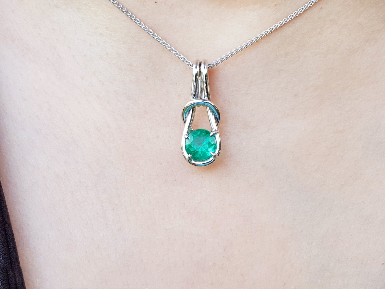 A white gold pendant with a round cut natural emerald on a chain, worn around the neck.