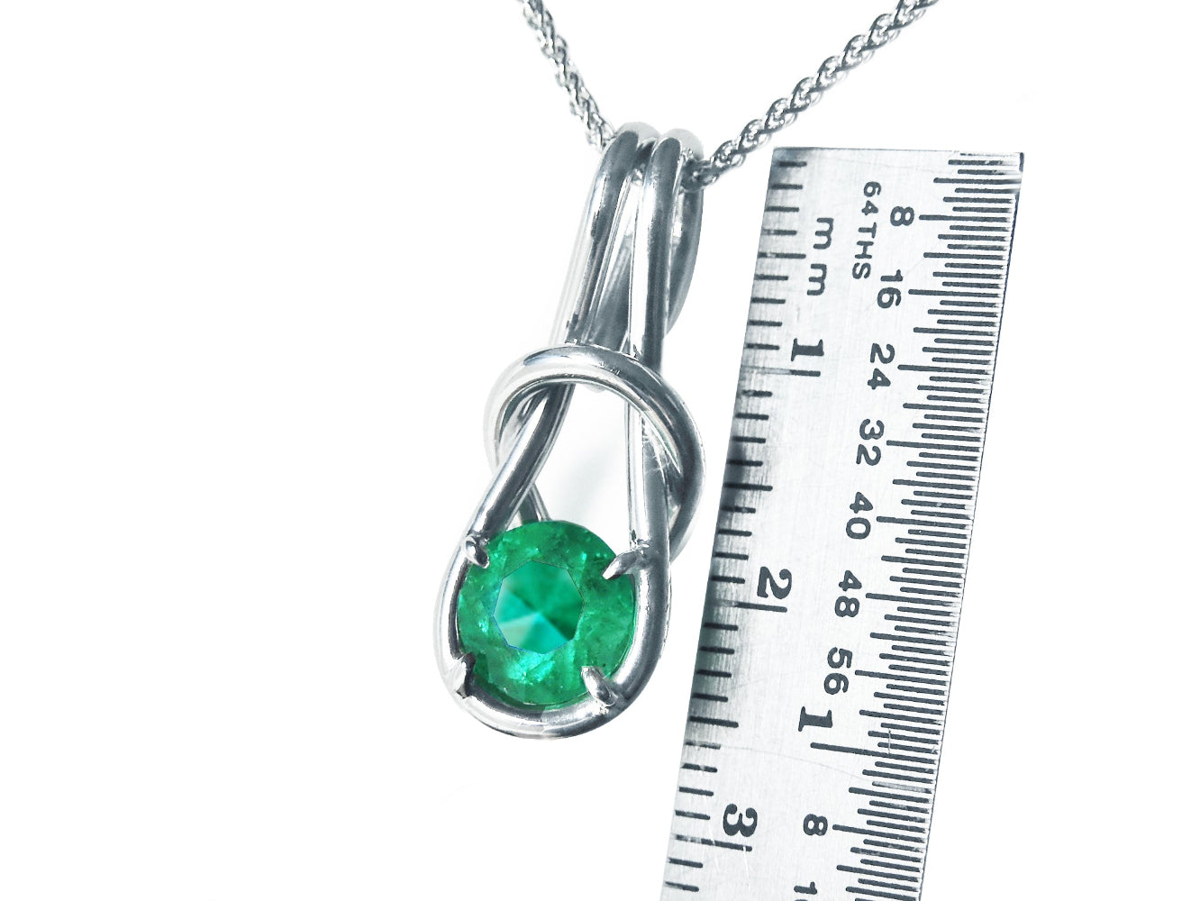 A white gold ;ove knot pendant with a round cut emerald next to a ruler for size reference on a white background.