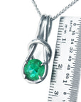 A white gold ;ove knot pendant with a round cut emerald next to a ruler for size reference on a white background.