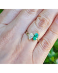 You and me emerald and diamond marquise ring shown worn on a person's finger against a blurred green background.