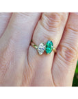 A 18k gold ring with a marquise-cut emerald and a diamond, shown worn on a person's finger against a blurred green background.