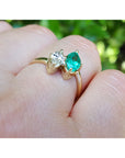 A gold ring with a marquise-cut emerald and diamond, worn on a person's finger with a blurred green outdoor background.