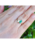 A 18k yellow gold ring toi et moi design with marquise emeralds and diamond shown worn on a person's finger with a blurred green garden background.
