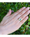 You and me emerald and diamond ring, worn on the ring finger of a hand, set against a background of green outdoor foliage.