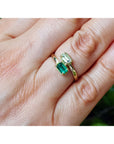 A close-up of a hand wearing two gold rings, one with a rectangular green emerald-cut gemstone and the other with a smaller round green stone.