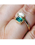A gold ring with a large emerald-cut green gemstone and a smaller clear rectangular gemstone, shown worn on a person's finger.