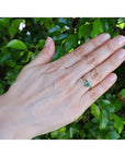 A gold ring toi at moi style with a natural emerald paired with a pear cut diamond worn on a person's ring finger, shown against a background of green foliage.