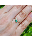A gold ring with a pear-cut emerald and a diamond, worn on a person's finger with a blurred green outdoor background.