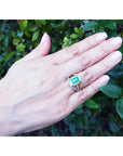 La mano de una mujer luciendo un anillo de oro amarillo con esmeralda sobre un fondo de follaje natural.