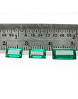 Three baguette-cut natural loose Colombian emeralds, deep bluish-green, displayed with a ruler for scale.