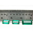 Three baguette-cut natural loose Colombian emeralds, deep bluish-green, displayed with a ruler for scale.