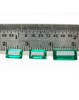 Three baguette-cut natural loose Colombian emeralds, deep bluish-green, displayed with a ruler for scale.