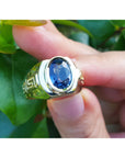 Men's genuine blue sapphire solitaire ring oval cut, top view held between fingers with leafy background