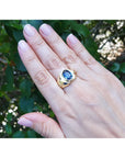 Hand wearing a gold blue sapphire solitaire ring with a blue gemstone against a green leafy background