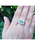 Hand wearing a men's gold ring with a colombian emerald against a leafy background.