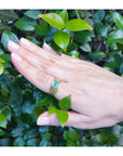 Hand wearing a gold pinky ring for men with a natural colombian emerald against a green leafy background.