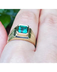 Gold ring for man with a bezel set emerald e on a finger against a blurred natural background.