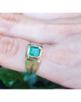 A yellow gold mens emerald pinky ring on a finger against a blurred natural background.