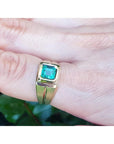 A yellow gold mens emerald pinky ring on a finger against a blurred natural background.