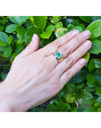 Hand wearing a gold emerald solitaire ring for men against a green leafy background.