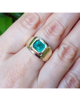 Gold ring with a bezel set genuine emerald on a hand against a blurred natural background.